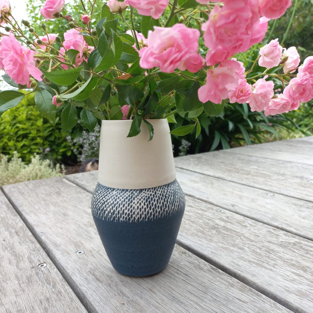 cette image montre un vase bicolore bleu et blan, sgraffité sur la partie bleue, avec des roses roses dedans posé sur une terrasse en bois