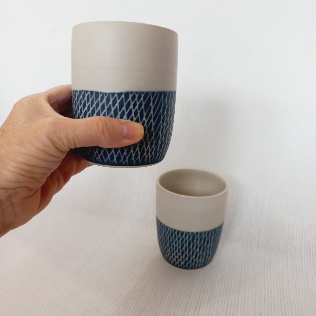 cette image montre un 1 tasse sans anse bicolores blanche et bleue et des motifs sur la partie colorée et un gobelet bleu posé à côté pour voir la différence de taille. je tiens la grosse tasse dans ma main