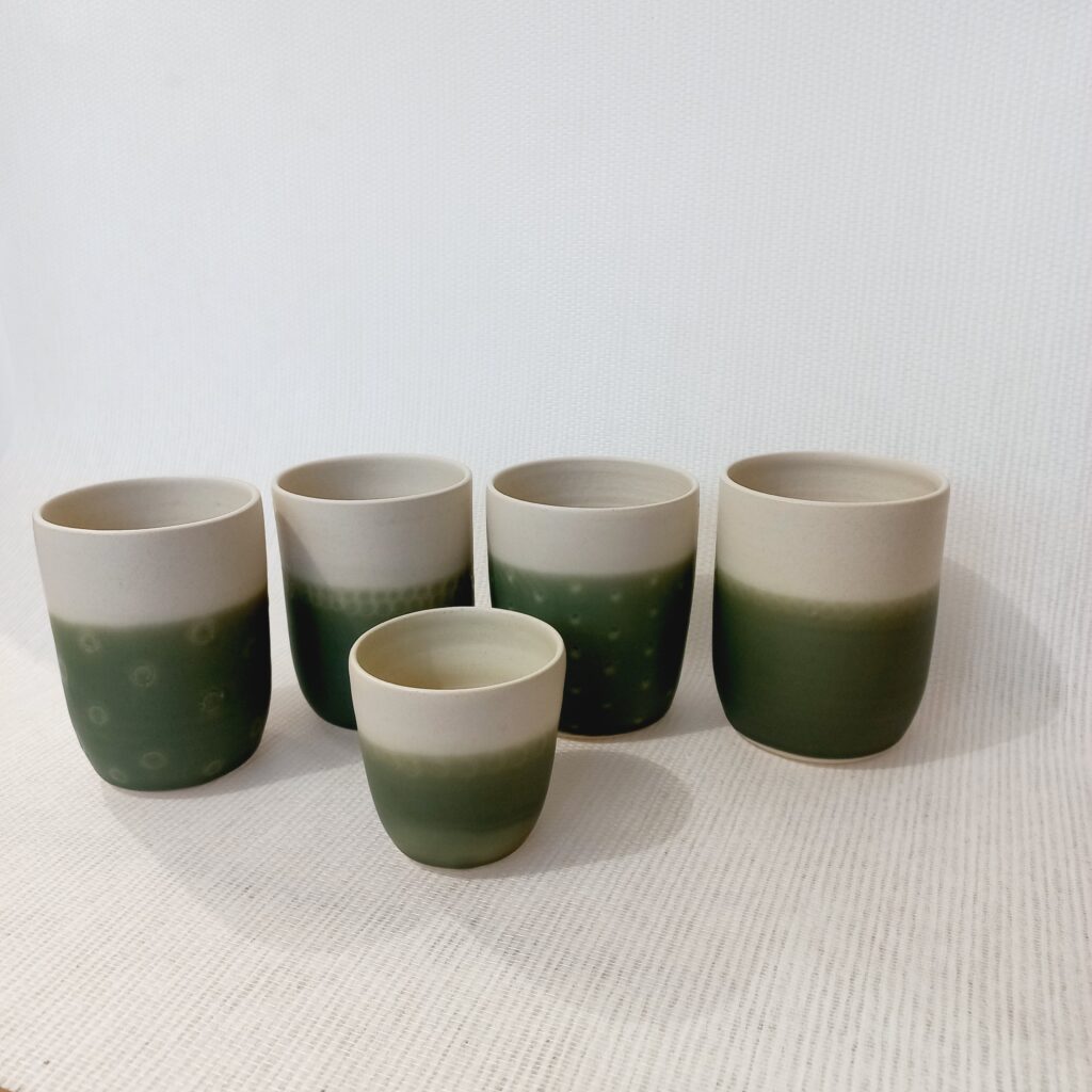 cette image montre un lot de 4 tasses sans anse bicolores blanche et verte et des motifs sur la partie colorée et un gobelet vert posé à côté pour voir la différence de taille. 