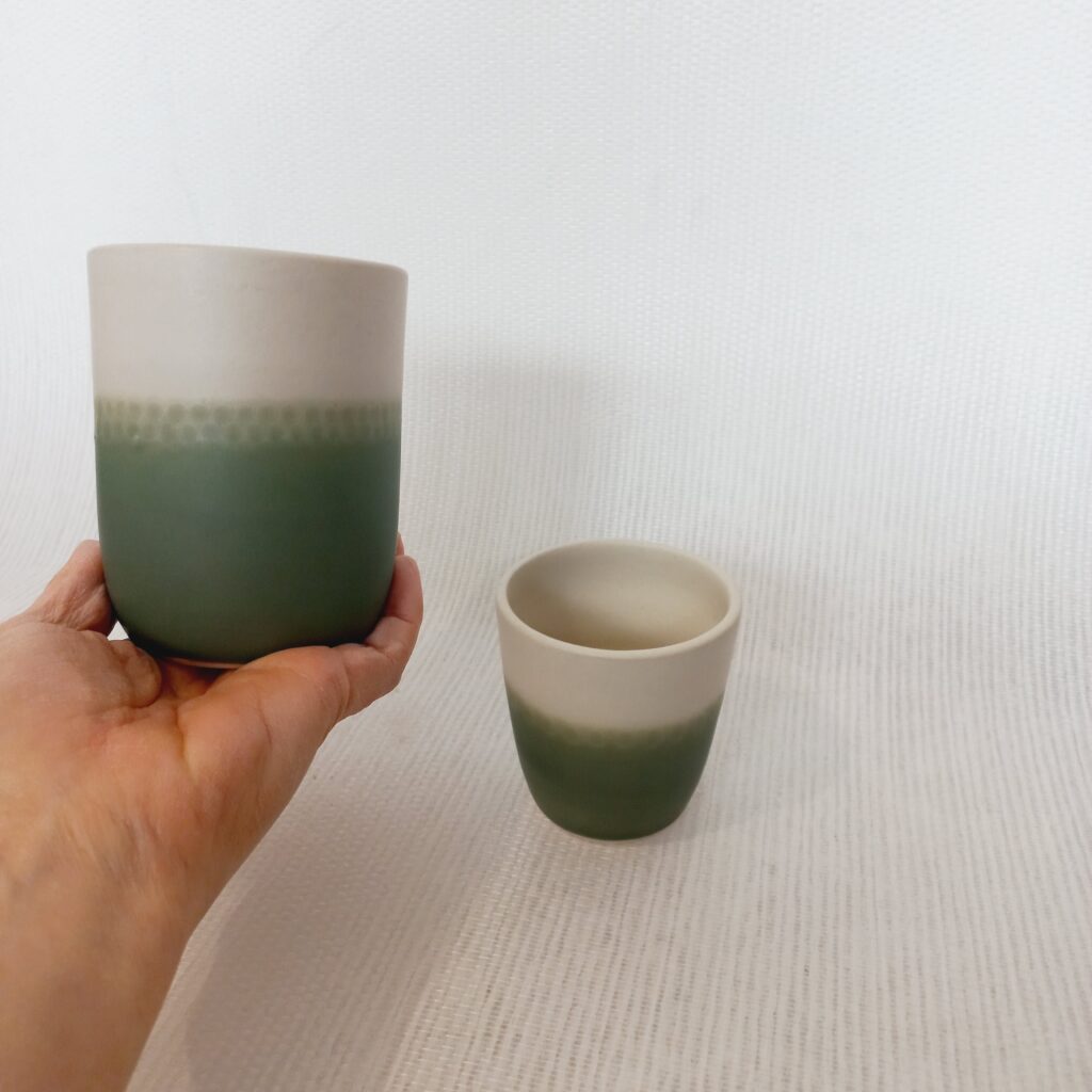 cette image montre 1 tasse sans anse bicolores blanche et verte et des motifs sur la partie colorée et un gobelet vert posé à côté pour voir la différence de taille. je tiens la grosse tasse dans ma main