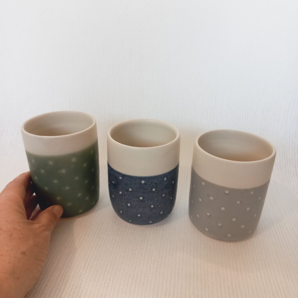 cette image montre un lot de 3 tasses sans anses bicolores blanhe verte bleue et grise etdes motifs à pois sur les parties colorées. ma main est sur la photo pour montrer la taille de la pièce